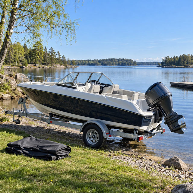 Bateau à moteur sur sa remorque au bord d'un lac calme au printemps, housse sombre pliée sur l'herbe à côté, moteur propre relevé