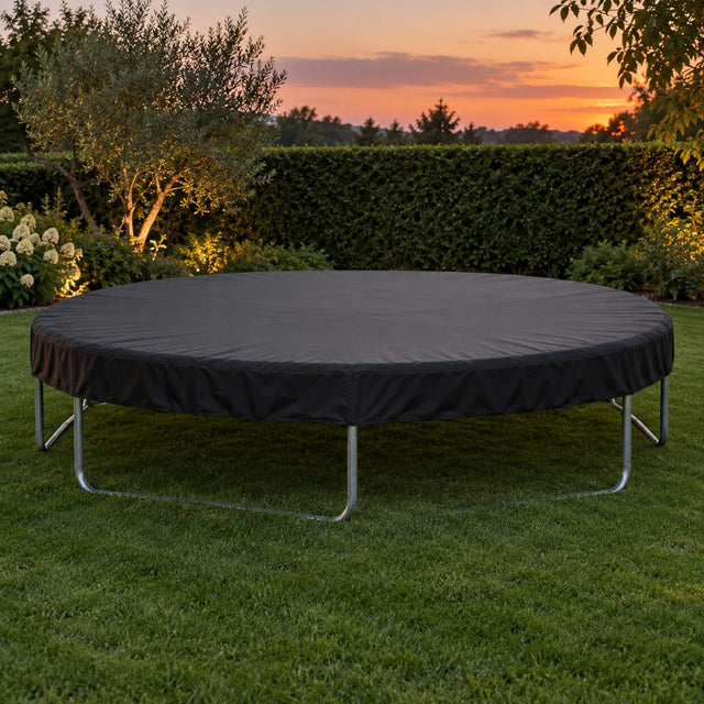Trampoline rond entièrement couvert d'une housse noire bien tendue dans un jardin au crépuscule, pelouse et haie derrière