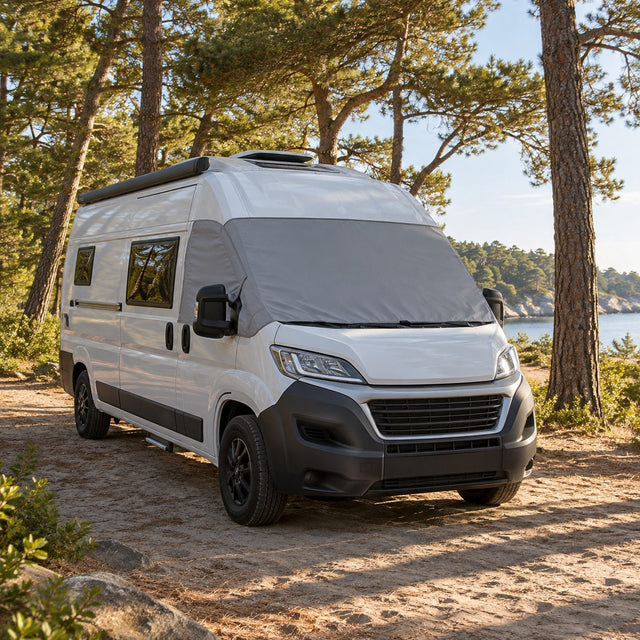 Camping-car blanc sous de grands pins un après-midi d'été, avec une housse de pare-brise grise en place et de longues ombres sur le sable