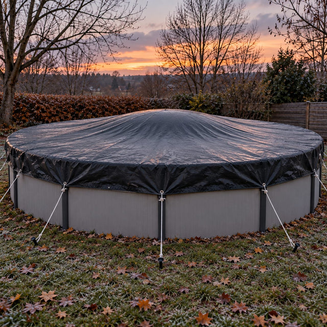 Piscine hors sol ronde sous une couverture d’hiver tendue avec une légère bosse au centre, cordes fixées à des piquets, à la tombée du jour